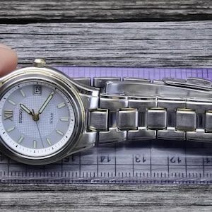 May include: A silver and gold tone Seiko Solar watch with a white face and black hands. The watch has a silver metal band with a fold-over clasp.