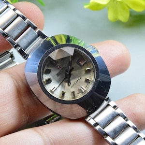 Könnte beinhalten: Eine Vintage-Armbanduhr in Silber mit einem geometrischen Zifferblatt und einem passenden Metallarmband. Das Zifferblatt der Uhr ist hell mit schwarzen Zeigern und Stundenmarkierungen. Die Uhr wird von einer Hand gehalten, die ihr Design und ihre Details zeigt.