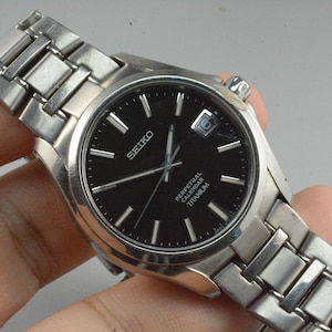 May include: A silver Seiko Perpetual Calendar Titanium watch with a black dial. The watch features silver hands and markers, a date window, and a silver link bracelet. The watch is a classic timepiece.