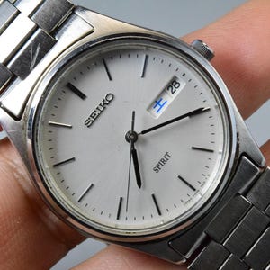 Op de afbeelding: Zilverkleurig Seiko Spirit horloge met een witte wijzerplaat, zwarte wijzers en een datumweergave. Het horloge heeft een zilveren metalen band. Het woord "SEIKO" is zichtbaar op de wijzerplaat. De datum toont het nummer 28.