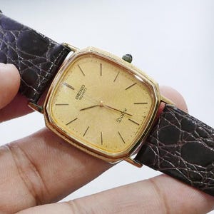 Op de afbeelding: Een goudkleurig Seiko Dolce quartz horloge met een vierkante wijzerplaat en een bruine leren band. De wijzerplaat heeft gouden wijzers en markeringen. De band is donkerbruin met een textuur, krokodilachtig patroon.