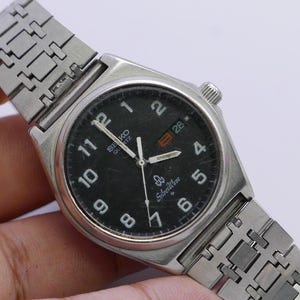 May include: A silver Seiko Quartz watch with a black face and silver hands. The watch has a silver metal strap and a date window at the 3 o'clock position.