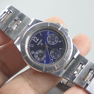Könnte beinhalten: Eine silberfarbene Seiko-Armbanduhr mit dunkelblauem Zifferblatt. Die Uhr hat ein Edelstahlarmband und ein rundes Zifferblatt mit weißen Ziffern und Zeigern. Sie verfügt über drei Hilfszifferblätter und eine Datumsanzeige. Die Uhr ist ein klassischer Zeitmesser.