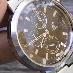 May include: A silver Citizen XC chronograph wristwatch with a black face and three subdials. The watch has a date window at the 3 o'clock position and the words "XC" and "CITIZEN" printed on the face.