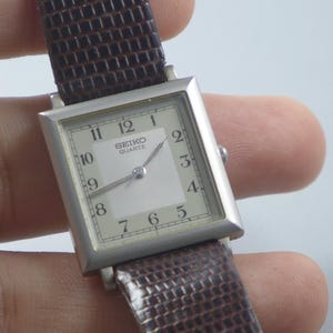 Puede incluir: Un reloj Seiko Quartz plateado con esfera cuadrada y correa de cuero marrón. La esfera es blanca con números y agujas negras. La correa tiene un patrón texturizado.