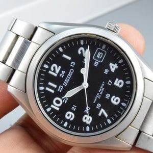 May include: A stainless steel Seiko wristwatch with a black dial, white Arabic numerals, and a date window at the three o'clock position. The watch features a metal link bracelet, luminous hands, and text on the dial reading Seiko, 23, and Water Resist 10 Bar.