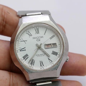 May include: A silver wristwatch with a white face and black Roman numeral hour markers. The watch has a date window and the words "Ricoh Quartz" printed on the face.