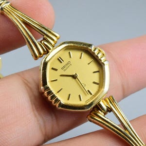 Pode incluir: Um relógio Seiko Quartz em tom dourado com caixa octogonal e mostrador dourado a condizer. O relógio apresenta um design de bracelete de elos múltiplos texturizados que se liga à caixa com um acessório metálico decorativo em leque.