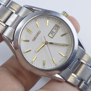 Könnte beinhalten: Silberfarbene Seiko-Uhr mit weißem Zifferblatt und goldfarbenen Stundenmarkierungen und Zeigern. Die Uhr zeigt das Datum und den Wochentag an. Die Uhr hat ein silberfarbenes Armband.