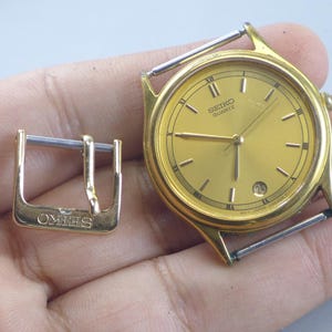 Könnte beinhalten: Eine goldfarbene Seiko Quartz-Uhr mit passender Schnalle. Die Uhr hat ein goldenes Zifferblatt mit schwarzen römischen Ziffern und Zeigern. Das Datum wird auf 3 Uhr angezeigt. Die Schnalle ist mit dem Wort "SEIKO" gestempelt.
