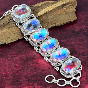 May include: A silver-toned bracelet featuring six large, iridescent, oval-cut gemstones. The gemstones display a rainbow of colors. The bracelet has an ornate, textured setting and a chain closure. The bracelet is displayed on a dark red surface.
