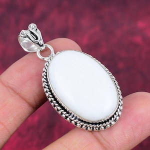 Puede incluir: Colgante blanco de forma ovalada con una fianza plateada y un borde decorativo. El colgante est&aacute; sujeto por una mano sobre un fondo rojo. El colgante es una joya.