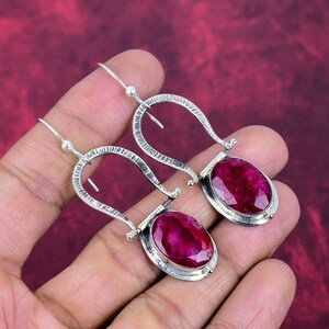May include: A pair of silver earrings featuring large, oval-shaped, deep red gemstones. The earrings have a unique, textured design with a hook closure. The gemstones are set in a silver bezel, creating a striking contrast.