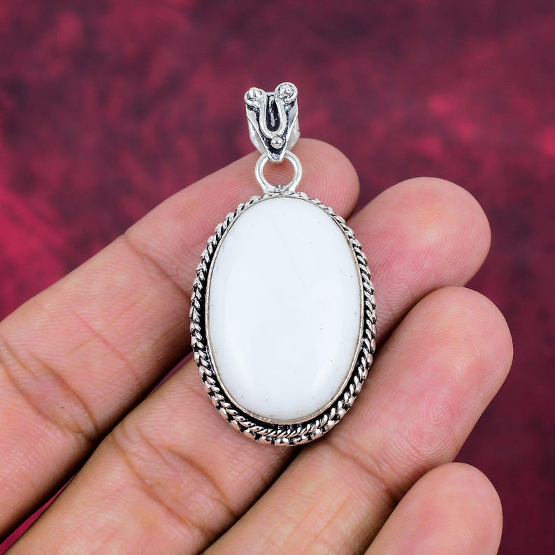 Puede incluir: Colgante ovalado de piedra blanca con marco y fianza de metal plateado. El colgante tiene un borde en forma de cuerda y una superficie lisa y pulida. La fianza tiene un dise&ntilde;o decorativo.