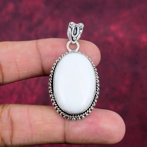 Puede incluir: Colgante ovalado de piedra blanca con marco y fianza de metal plateado. El colgante tiene un borde en forma de cuerda y una fianza decorativa. La piedra es lisa y pulida, sobre un fondo rojo oscuro.