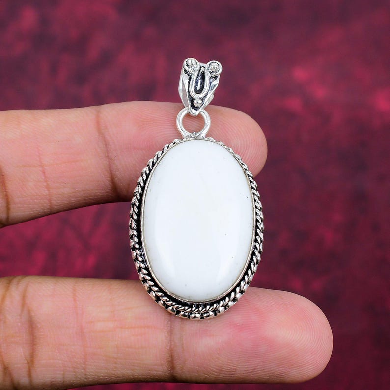 Puede incluir: Colgante ovalado de piedra blanca con marco y fianza de metal plateado. El colgante tiene un borde en forma de cuerda y una fianza decorativa. La piedra es lisa y pulida, sobre un fondo rojo oscuro.