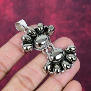 Puede incluir: Un colgante de plata con un grupo de piedras pulidas de aspecto metálico. El colgante tiene una piedra central ovalada y piedras más pequeñas en forma de lágrima que lo rodean. El colgante está sobre un fondo rojo.