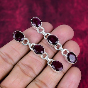 May include: A pair of silver earrings featuring three oval, deep red gemstones linked together. The earrings have a simple, elegant design with a polished finish. The gemstones are set in a bezel setting, and the earrings are shown against a dark red background.