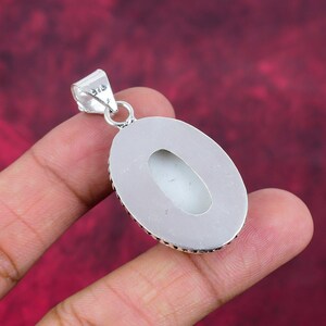 Puede incluir: Un colgante plateado con una piedra ovalada de color claro. El colgante tiene un borde detallado y texturizado y un peque&ntilde;o lazo para una cadena. La piedra tiene una superficie lisa y pulida. El colgante est&aacute; sostenido por una mano sobre un fondo rojo.
