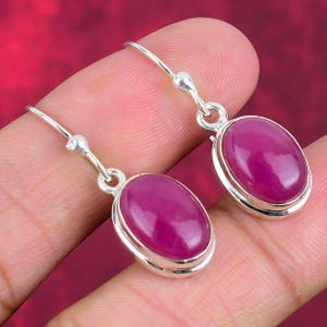 May include: A pair of silver drop earrings featuring oval, magenta-colored gemstones. The gemstones are set in a silver bezel and have a polished, smooth surface. The earrings are displayed against a red background.