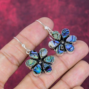May include: A pair of flower-shaped earrings with iridescent abalone shell petals and silver-tone metalwork. The earrings have a hook closure and a dark center, showcasing a vibrant mix of blues, greens, and purples.