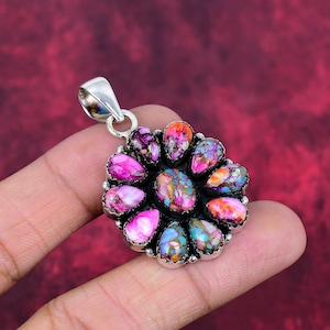 May include: A silver pendant with a flower-like design. The pendant features a central oval stone surrounded by teardrop-shaped stones in shades of pink, purple, orange, and blue. The stones have a speckled appearance and are set in a silver metal.