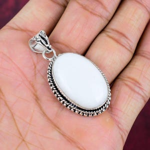 Puede incluir: Un colgante de plata con una piedra ovalada blanca. La piedra est&aacute; rodeada por un borde plateado trenzado detallado. El colgante tiene una fianza decorativa en la parte superior para una cadena. La joya se muestra en una mano.