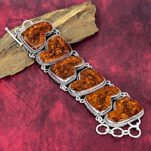 May include: A silver bracelet featuring six amber stones set in a heart shape. The stones are a deep orange colour and have a natural, organic look. The bracelet is made of a chain link style and has a clasp closure.