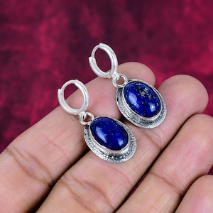 Puede incluir: Pendientes de aro plateados con piedras ovaladas de lapislázuli azul oscuro. Las piedras tienen un aspecto moteado y están engastadas en un marco plateado texturizado. Los pendientes se muestran sobre un fondo rojo.