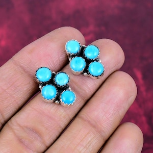 May include: A pair of turquoise stud earrings. Each earring has five round, bright blue turquoise stones set in a silver-coloured metal. The earrings are shown against a blurred, reddish-brown background.