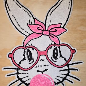 May include: A white bunny with pink heart-shaped glasses and a pink bow, blowing a pink bubble. The bunny has black eyes and a black nose.
