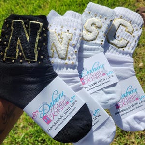 May include: Three pairs of white and black socks with the letters N, S, and C embroidered on them. The socks are decorated with gold glitter and white beads. The socks are packaged with a white label that reads "Dabrene Kiddie Kloset Handmade With Love 90% Cotton 5% poly 5% Elastic".
