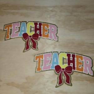 May include: Two colorful glitter "TEACHER" signs with red bows. The signs are made of felt and have a gold glitter outline.