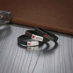 May include: Three braided leather bracelets with metal medical alert tags. One tag is silver with a red medical symbol and the text Hypertention Blood thinner. Another is black with a red symbol. The third is silver with a red symbol.