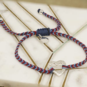 May include: A braided cord bracelet with a silver heart charm. The cord is red, blue, and gray.