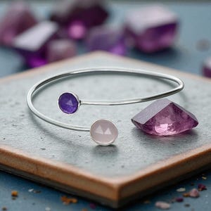 Peut inclure: Un bracelet argenté orné d'une pierre précieuse violette et d'une rose. Le bracelet ouvert est présenté avec un cristal violet facetté et d'autres cristaux violets en arrière-plan. Bijoux.