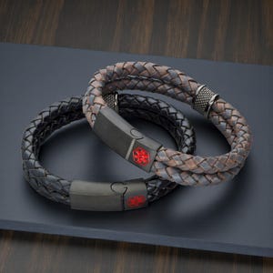 May include: Two braided leather bracelets. One is black, the other is brown and gray. Each bracelet has a black clasp with a red medical symbol. The bracelets are on a dark surface.