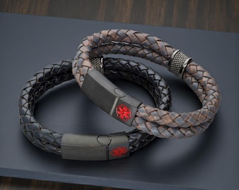 May include: Two braided leather bracelets. One is black, the other is brown and grey. Each bracelet has a black clasp with a red medical symbol. The bracelets are on a dark surface.