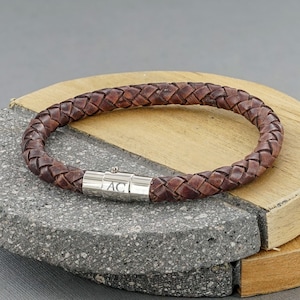 Op de afbeelding: Een bruin leren armband met een zilveren magneetsluiting. De sluiting is gegraveerd met de initialen "AC".