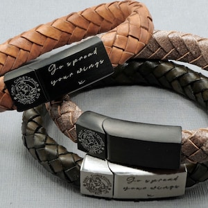 May include: Two leather bracelets with black metal clasps. One bracelet is brown and the other is green. Both bracelets have a silver charm with the words "Saint Christopher Protect Us" engraved on them. The clasps have engraved messages: "Go spread your wings" and "Go spread your wings".