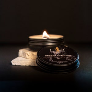 Op de afbeelding: Een zwarte kaarsblik met de tekst "HOUSE OF GEB" en "DESERT CAMPFIRE" gedrukt op het etiket. De kaars is aangestoken en de was smelt. De kaars staat op een witte steen.