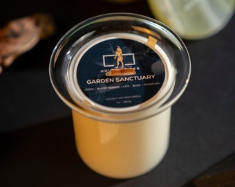May include: A white candle with a black and gold label that reads "HOUSE OF EB GARDEN SANCTUARY LEMON | BLOOD ORANGE | LIME | BASIL | ROSEMARY" and a small illustration of a person. The candle is in a clear glass jar with a lid.