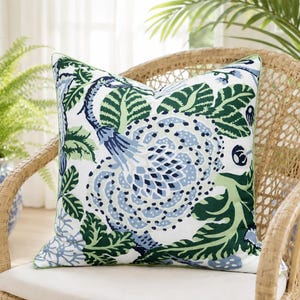 May include: A square throw cushion featuring a white background with a large, intricate floral pattern in shades of dark green, light green, and various tones of blue. The design includes stylised leaves and blossoms with a piped green edge.