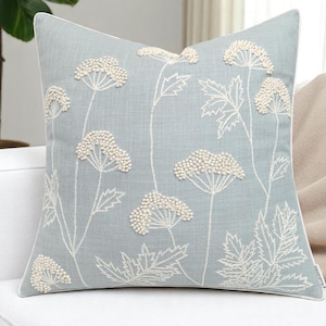 Op de afbeelding: Een vierkant decoratief kussen met een lichtblauwe achtergrond en witte bloemborduurwerk. Het kussen heeft een witte rand en is versierd met witte bladeren en bloemhoofden met textuur en kralen. Het ontwerp is bloemig.