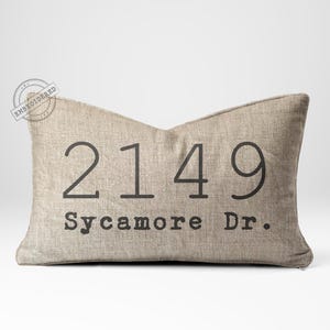 May include: A rectangular beige pillow with the address "2149 Sycamore Dr." printed in dark gray. The pillow has a textured appearance and a decorative shape. The word "EMBROIDERED" is stamped on the top left corner.