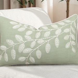 Puede incluir: Un cojín rectangular verde salvia con un diseño de hojas bordadas en blanco. El cojín tiene un borde blanco y está colocado sobre un sofá blanco. Un elemento decorativo para el hogar.