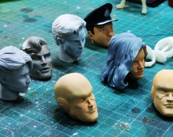 May include: A collection of unpainted plastic heads for model making. The heads are in various stages of completion, with some having hair and others being bald. The heads are all different colors, including white, gray, brown, and blue.