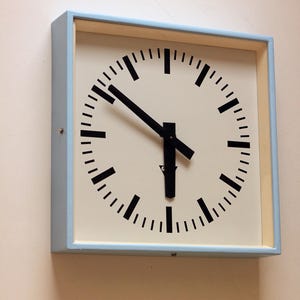 May include: A square wall clock with a light blue frame and an off-white face. The clock features black hour and minute hands, along with black tick marks for each hour and minute interval. The hands are positioned to indicate approximately 5:20.