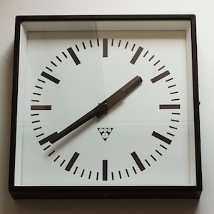 Può includere: Orologio da parete quadrato con quadrante bianco e lancette e segni neri. L'orologio ha una cornice nera e un piccolo logo triangolare in basso al centro. Le lancette dell'orologio indicano circa le 2:30.