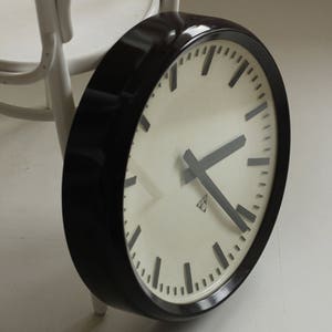 May include: A large, round clock with a black frame and a cream-colored face. The clock has black hands and markers, and is propped against a white chair. The clock's diameter is approximately 12 inches.